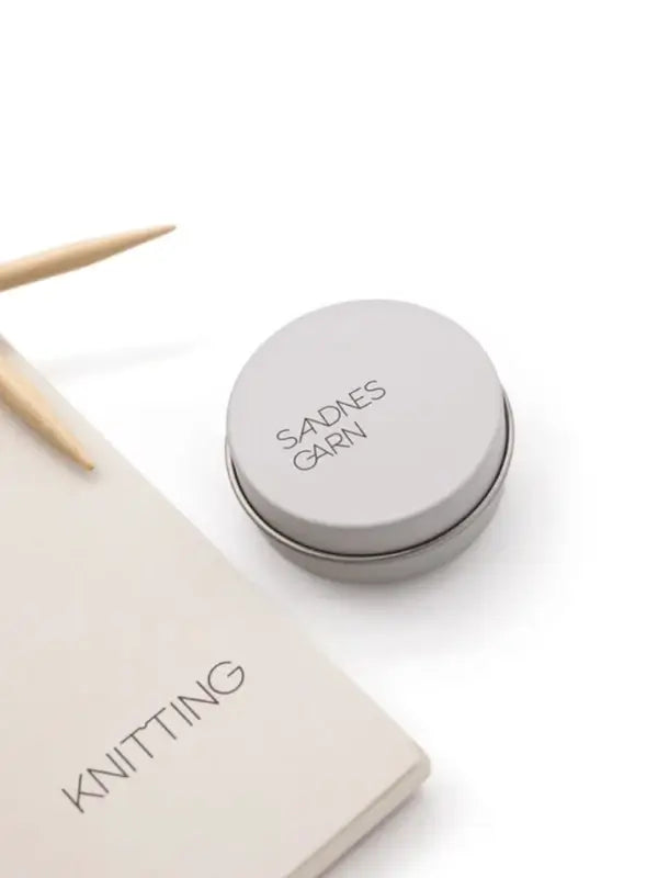 A round tin labeled Sandnes Garn Silmukkamerkit sits on a white surface beside a beige KNITTING notebook and two light wooden knitting needles.