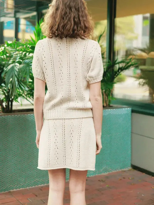 Model wears the Cornelia Tee & Skirt neuleohje ENG with Sandnes Garn Line, standing outdoors against green plants and blue-tiled walls, their shoulder-length curls visible from behind.