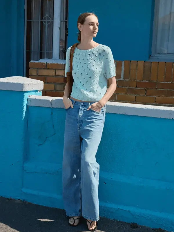 Someone stands outdoors by a blue wall, wearing the Elodie Tee neuleohje ENG knitted from Sandnes Garn Line, paired with loose-fitting jeans, sandals, and a brown shoulder bag. A window and exposed brick appear in the background.
