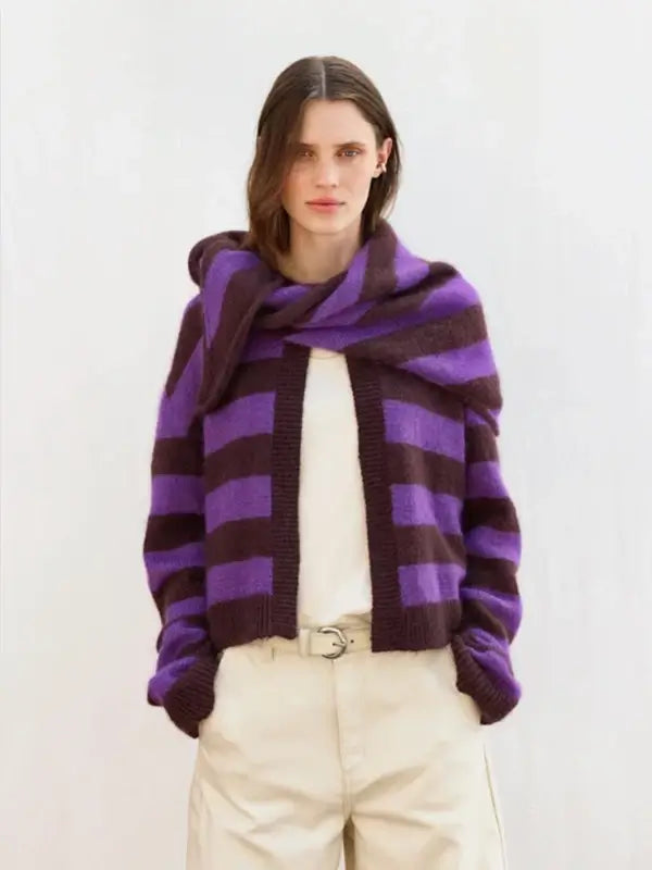 A person wearing a purple and dark brown striped sweater with the Sandnes Garn 2606-06 DIY Mango Scarf – neuleohje suomeksi draped over their shoulders, paired with beige pants, stands against a plain light background.