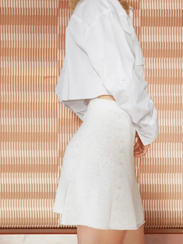 A person in a long-sleeve white cropped shirt and high-waisted white knit skirt, styled as a chic set inspired by the Milly Sweater & Skirt Neuleohje Suomi +Sandnes Garn line, stands in profile before a striped beige and brown background.