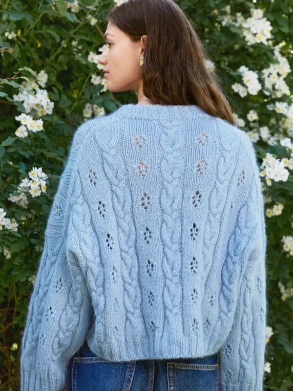 A person with long brown hair, wearing a light blue Sandnes Garn 2606-11 Junia Sweater – neuleohje suomeksi PDF and blue jeans, stands outdoors in front of a bush with white flowers.