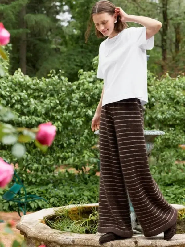 A person wearing a white t-shirt and wide, brown striped pants stands on the edge of a stone fountain in a garden with blooming pink roses and greenery, holding a copy of the Sandnes Garn Riley Sweater & Riley Pants – neuleohje suomeksi, PDF.