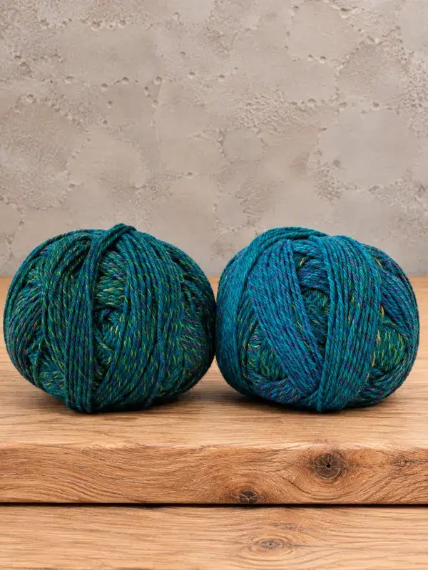 Two balls of Schoppel-Wolle Edition 3 yarn in variegated teal, blue, and yellow sit side by side on a wooden surface.