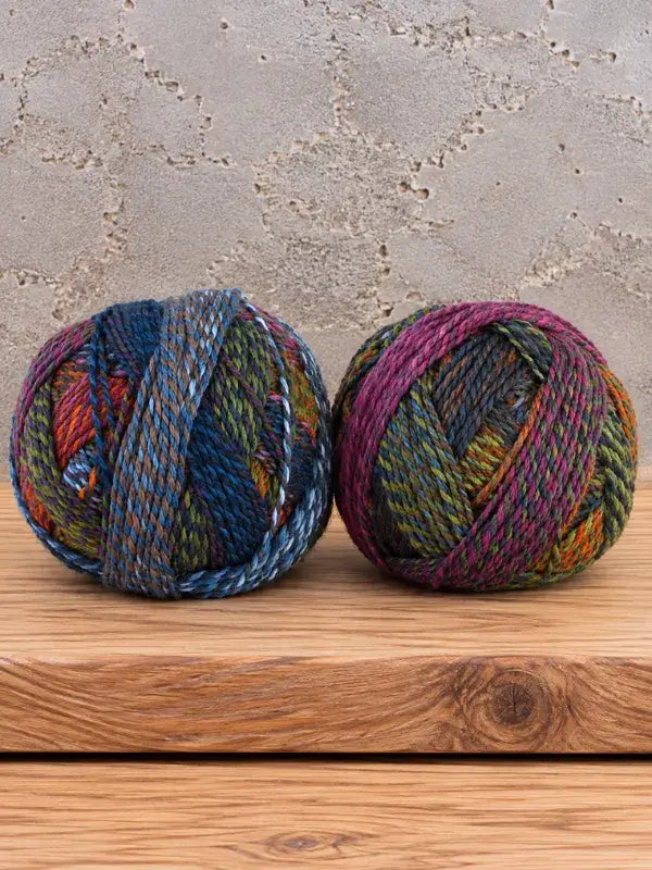 Two balls of Schoppel-Wolle Edition 3 variegated yarn in shades of blue, green, and magenta sitting on a wooden surface.