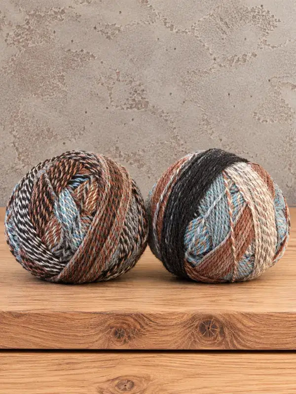 Two balls of Schoppel-Wolle Edition 3 variegated yarn in shades of brown, tan, blue, and black sitting on a wooden surface.