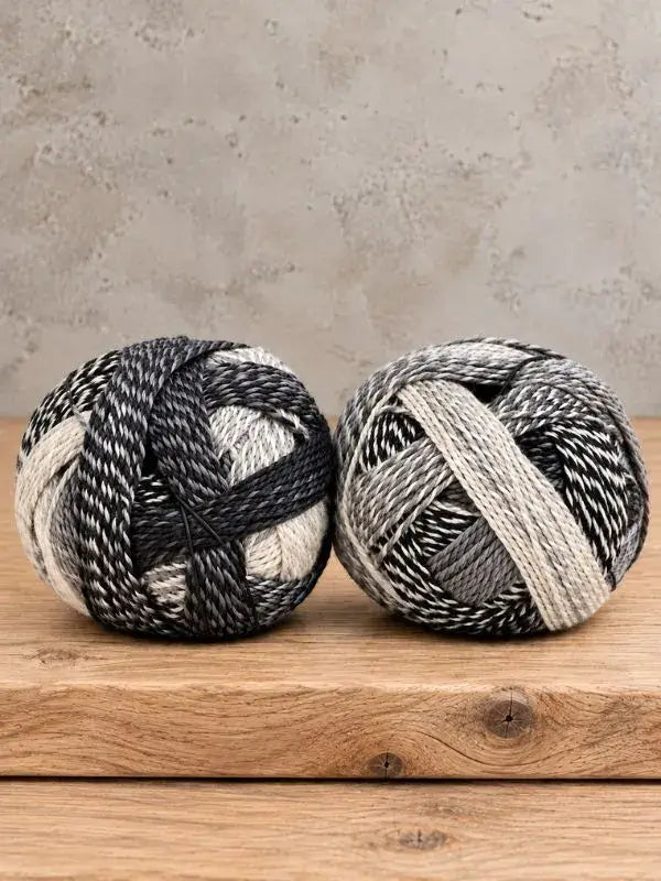 Two balls of Schoppel-Wolle Edition 3 variegated yarn in shades of black, grey, and white sitting on a wooden surface.