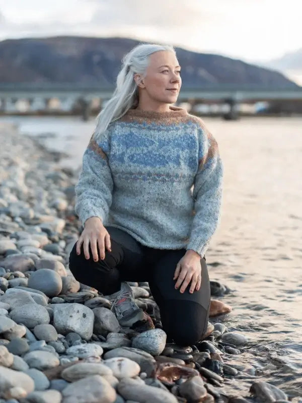 A woman with long white hair kneels on a rocky shore, wearing the Sisu Knitwear Lohineule neuleohje Suomi PDF sweater and black pants, gazing thoughtfully at mountains and shimmering water in the background.