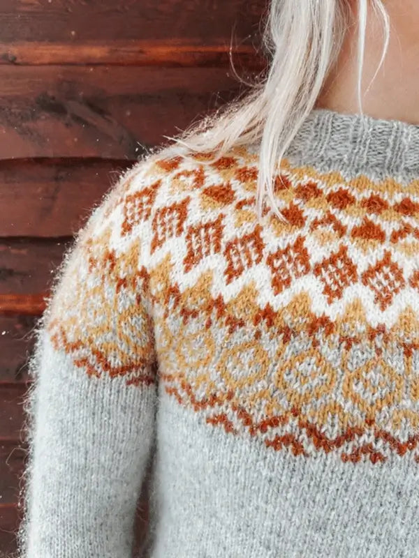 A person with long, light hair wears a gray Sisu Knitwear Mikkelineule sweater, showcasing a Nordic-inspired geometric pattern in brown, gold, and white across the shoulders and chest.