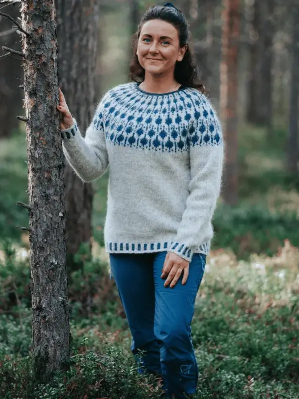 A woman stands in a forest, smiling with one hand on a tree trunk. She wears the Sisu Knitwear Mustikkaneule neuleohje Suomi PDF knit sweater with a white base and blue patterned yoke, surrounded by lush greenery and tall trees.