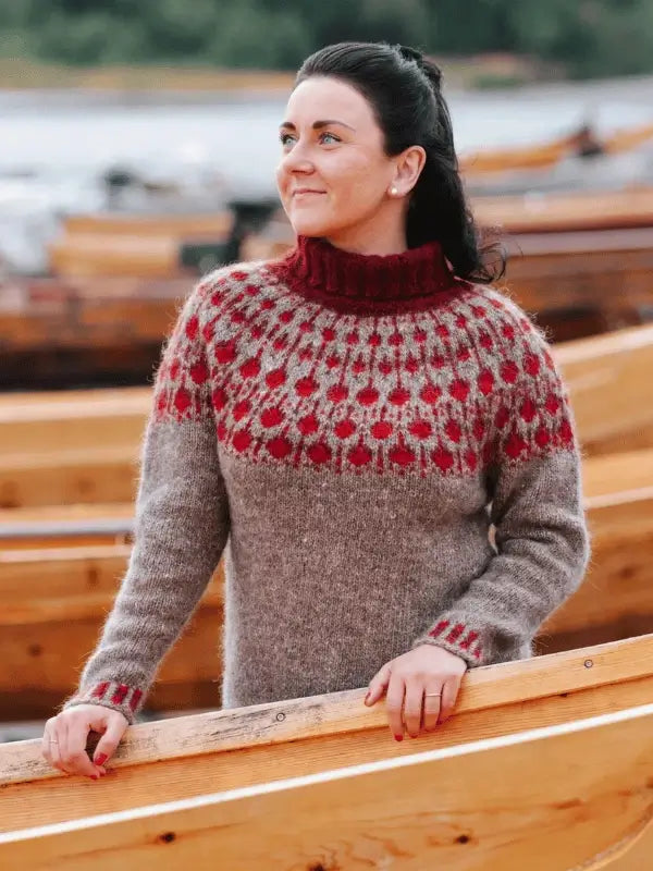 A woman with dark hair in a ponytail stands among wooden boats, wearing the Sisu Knitwear Puolukkaneule neuleohje Suomi PDF sweater with gray and red patterns. She smiles slightly as calm water and trees create a peaceful backdrop.