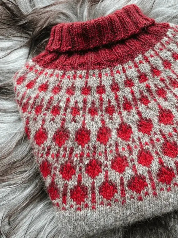 The Sisu Knitwear Puolukkaneule neuleohje Suomi PDF features a folded women’s sweater with a high collar and a red-gray Nordic geometric pattern, displayed on a soft gray surface.