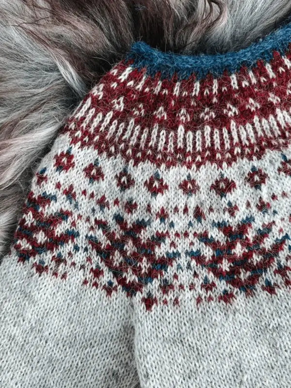 A close-up of the Sisu Knitwear Rovaneule neuleohje Suomi PDF sweater, featuring a traditional Nordic pattern in burgundy, blue, and white, lying on gray and white fur.
