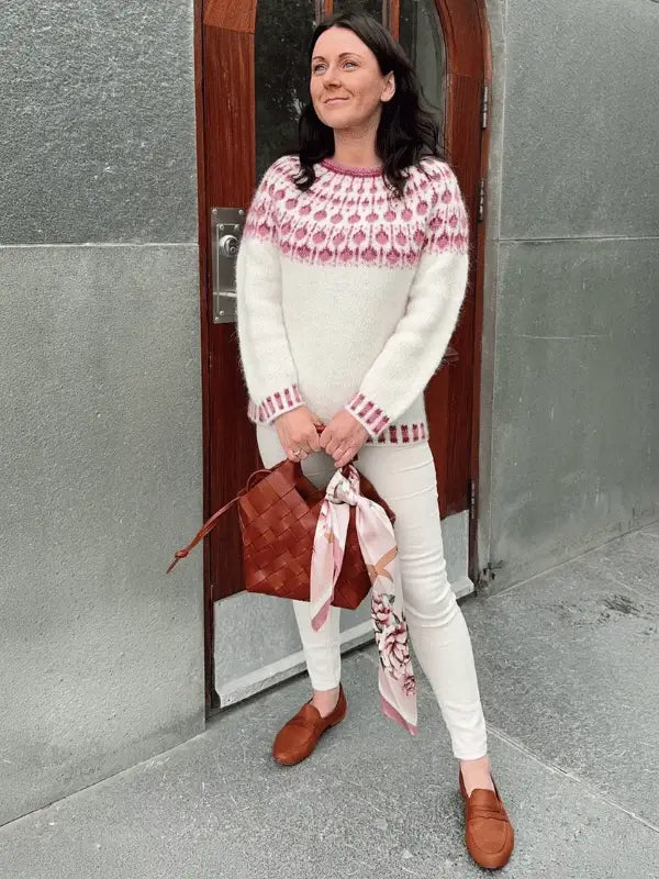 A woman stands before a wooden door wearing a white Sisu Knitwear Vadelmaneule neuleohje Suomii sweater with a pink pattern, white pants, brown loafers, and holding a brown woven bag adorned with a pink scarf.