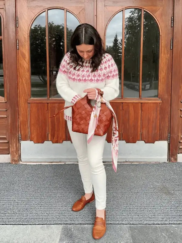 A woman stands on a mat by wooden doors, wearing white pants, brown shoes, and a Sisu Knitwear Vadelmaneule neuleohje Suomen PDF sweater with pink patterns. She holds a brown woven bag with a pink scarf tied to it and looks down.