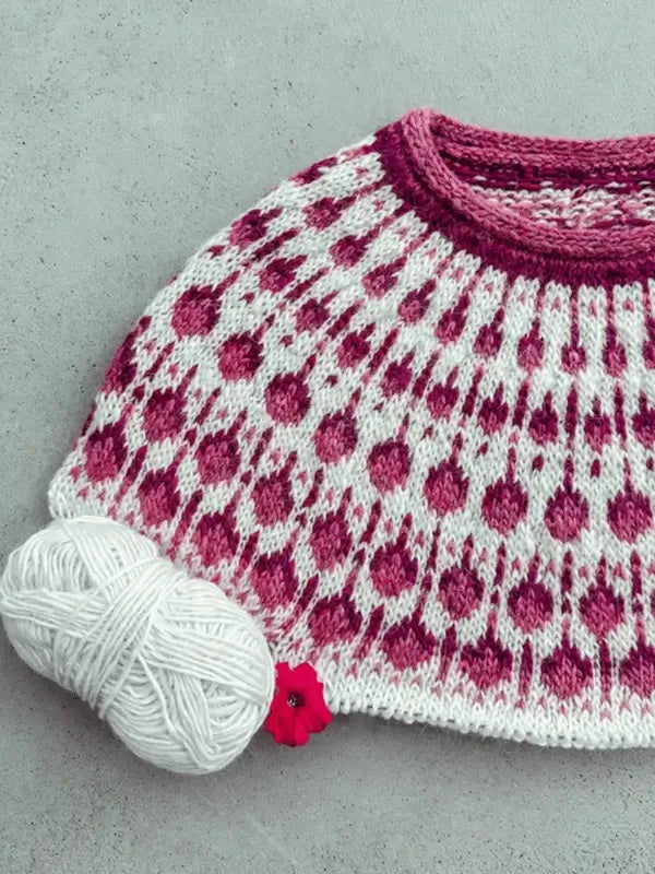 A round yoke sweater in white and magenta Lettlopi is partially knitted. A ball of white yarn and a red flower stitch marker sit on the Sisu Knitwear Vadelmaneule neuleohje Suomii PDF, against a plain gray background.