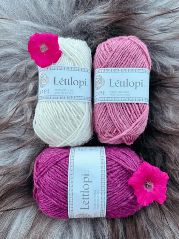 Three skeins of yarn in cream, pink, and magenta with bright pink flowers rest on a furry surface—ideal inspiration for your next Sisu Knitwear Vadelmaneule neuleohje Suomii PDF project.