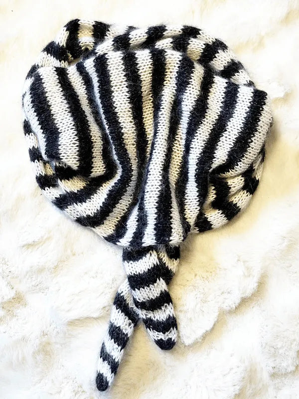 The Somany huivi ohje PDF features a black and white striped knitted blanket, crafted with Knitting for Olive Merino, displayed on a fluffy white surface in a stingray-like shape with two elongated tail sections.