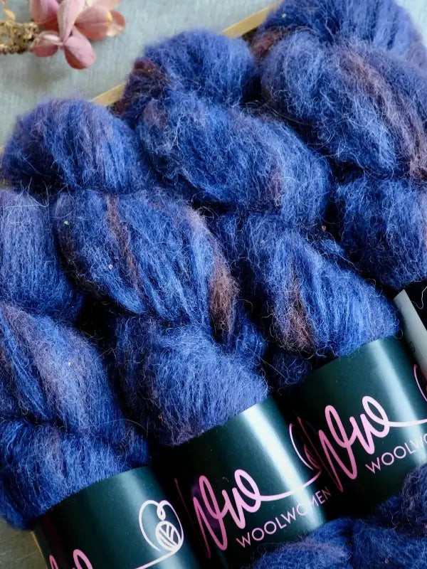 Three skeins of deep blue, fluffy käsinvärjätty WoolWomen Alpaca Glimmer yarn are arranged side by side, with a small pink flower and a twig visible in the corner.
