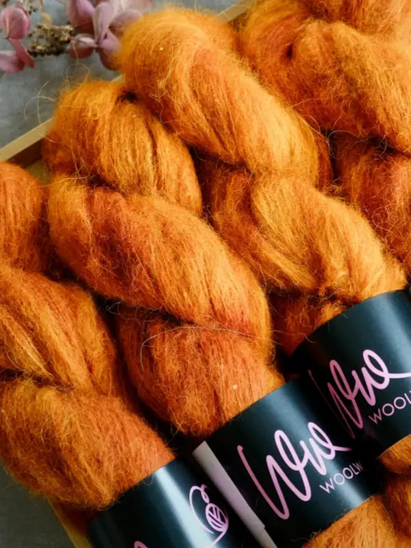 Three skeins of WoolWomen Alpaca Glimmer—fluffy, orange baby alpaca yarn—are neatly arranged in a box, each wrapped with a black label featuring pink writing. Pink flowers can be seen in the background.