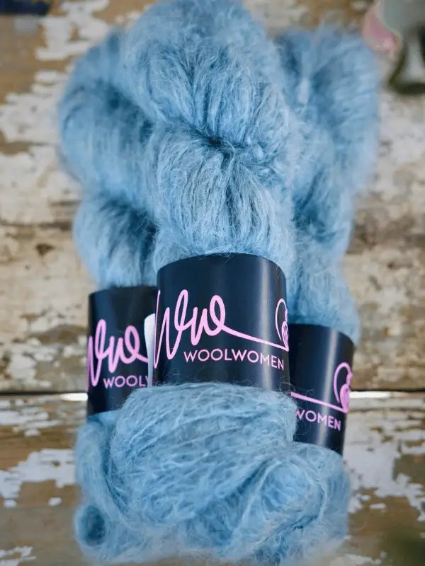 Three skeins of light blue WoolWomen Fluff Mohair yarn with black and pink bands rest on a rustic wooden surface—a käsinvärjätty lanka ideal for cozy projects.