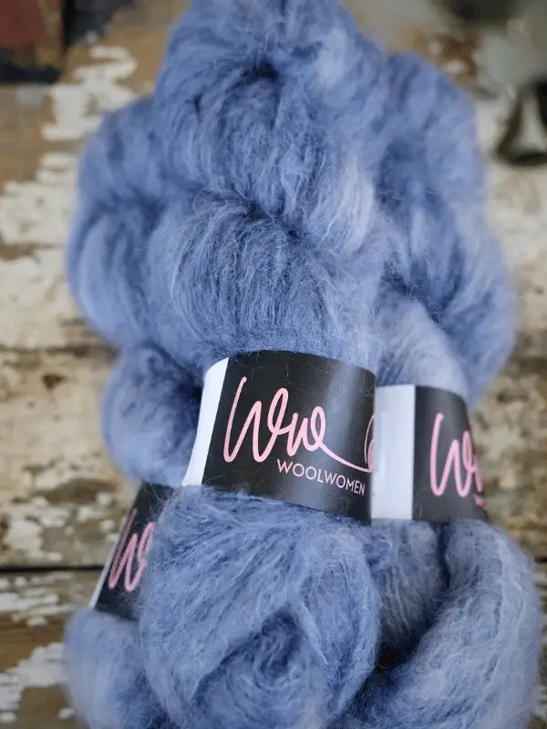 Two bundles of soft, fluffy blue WoolWomen Fluff Mohair yarn with black and pink labels are placed on a rustic, weathered wooden surface.