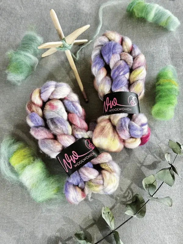 Two skeins of WoolWomen Kehruuvilla BFL Käsinvärjätyt, a wooden drop spindle, green and yellow loose fiber, and dried eucalyptus are arranged on a grey surface for spinning.
