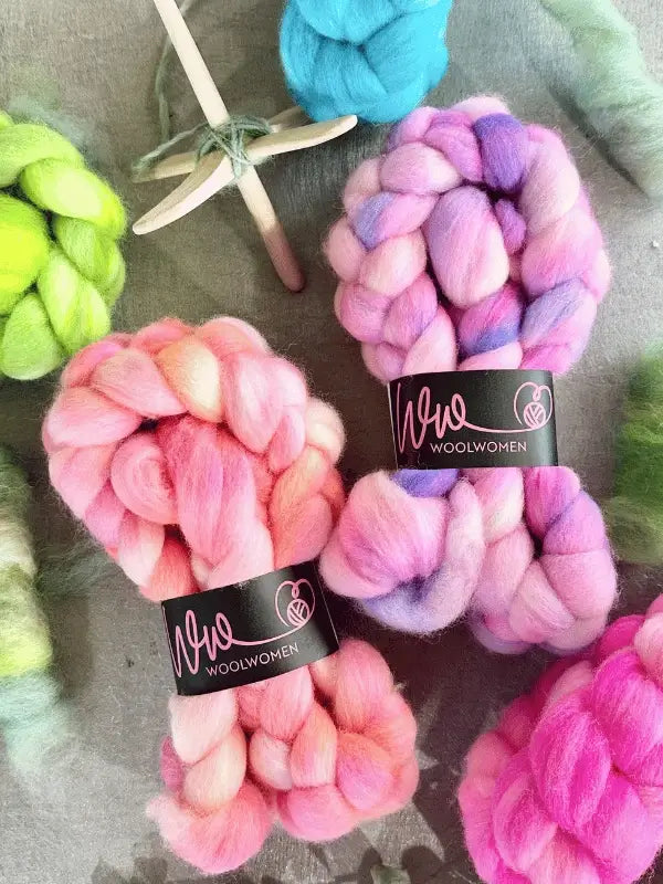 Colorful bundles of WoolWomen kehruukuitu Corriedale käsinvärjätty in pink, purple, and green are on a gray surface. A wooden drop spindle with teal fiber is present, and two bundles have WoolWomen labels.