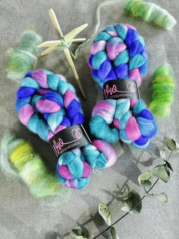 Two colorful braids of WoolWomen kehruukuitu Polwarth käsinvärjätty in blue, pink, and purple rest on a grey surface near a wooden spindle, green Polwarth fiber tufts, and dried leaves—perfect for your kehrääminen projects.