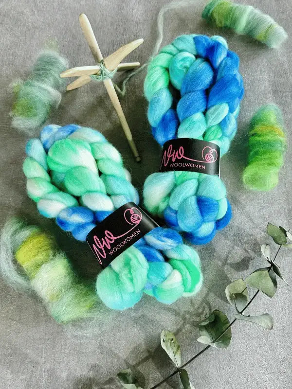 Two braids of blue and green WoolWomen kehruukuitu Polwarth käsinvärjätty lie on a gray surface, surrounded by carded wool, a wooden spinning spindle, and dried eucalyptus sprigs.