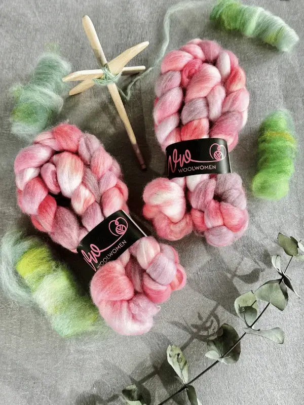 Two bundles of WoolWomen kehruukuitu Polwarth käsinvärjätty in pink and purple rest on a gray surface, surrounded by green fiber, a wooden drop spindle for spinning, and eucalyptus branches.