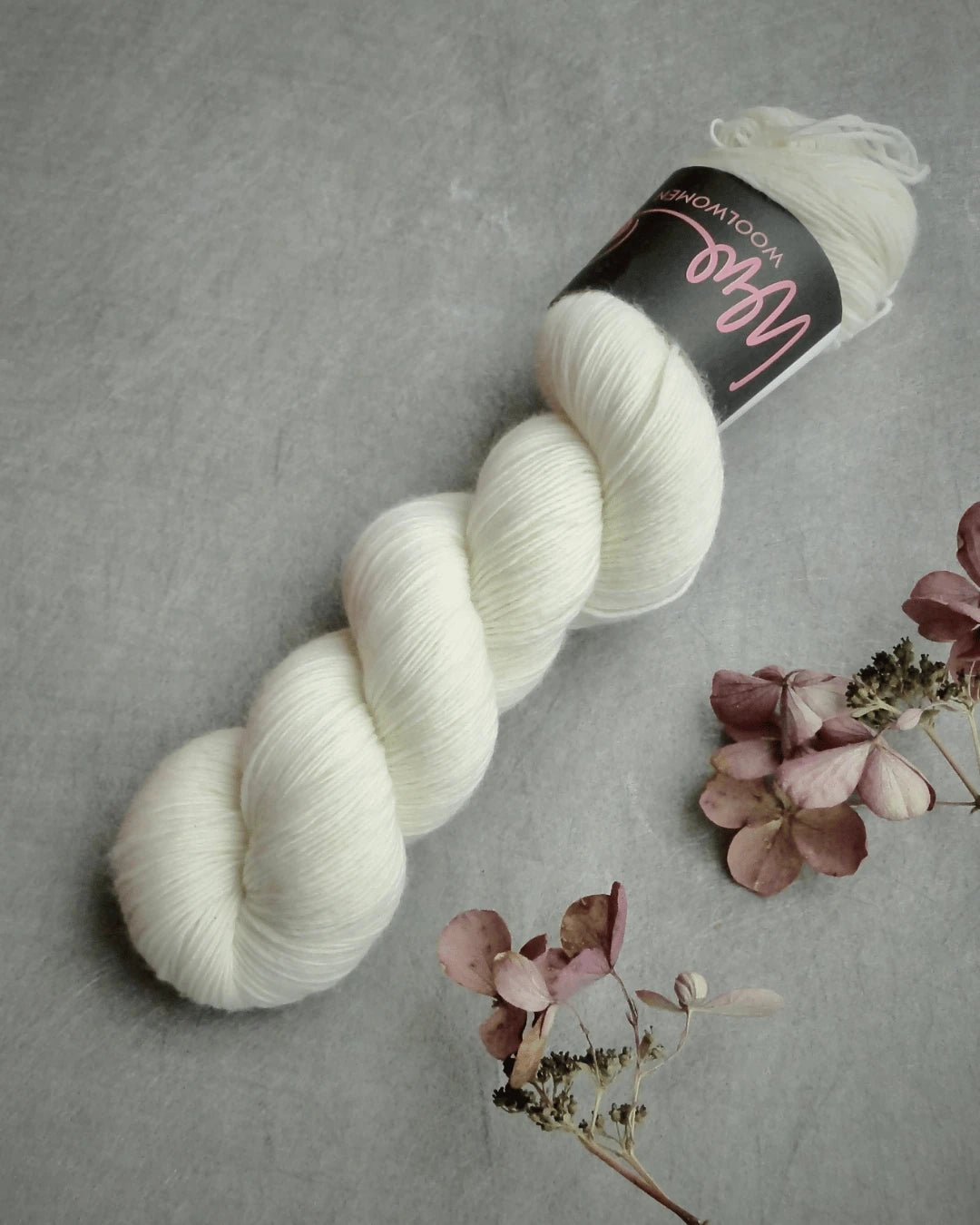 A twisted skein of WoolWomen Merino Single easywash-merino yarn with a black label rests on a gray surface beside a few dried pink and brown flowers.