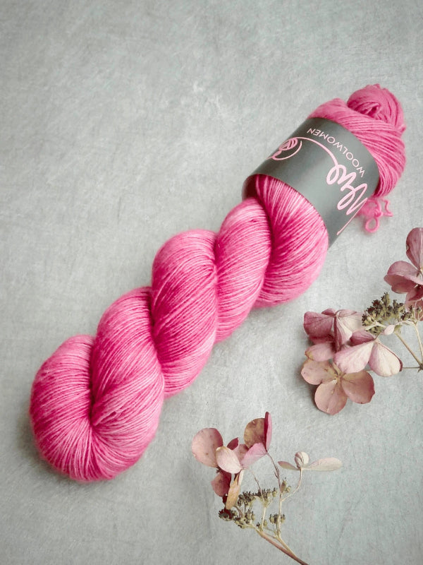 A skein of WoolWomen Merino Single käsinvärjätty merinovilla yarn in bright pink, with its label, rests on a light gray surface beside dried hydrangea flowers in soft brown and pink hues.