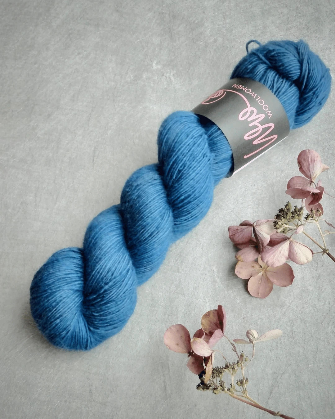 A skein of WoolWomen Merino Single yarn rests on a light gray surface beside several dried pink and beige hydrangea flowers.
