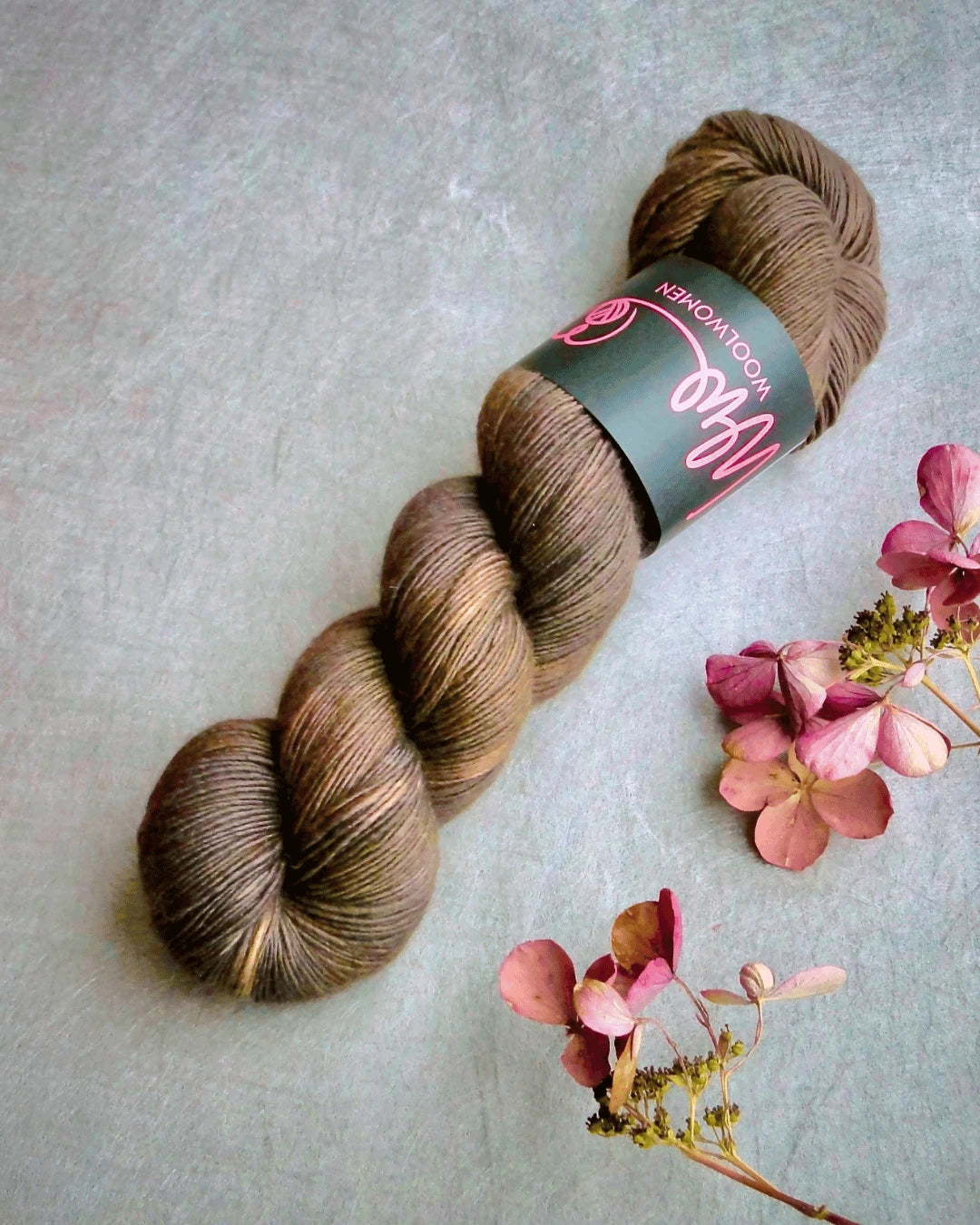 A skein of WoolWomen Merino Single yarn in brown, with a black and pink label, rests on a gray surface beside pink dried hydrangea flowers.