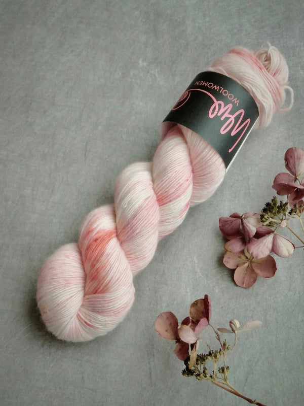 A skein of WoolWomen Merino Single, pink and white variegated käsinvärjätty merinovilla yarn, rests on a gray surface beside dried pink hydrangeas. The skein features a black label with pink writing.