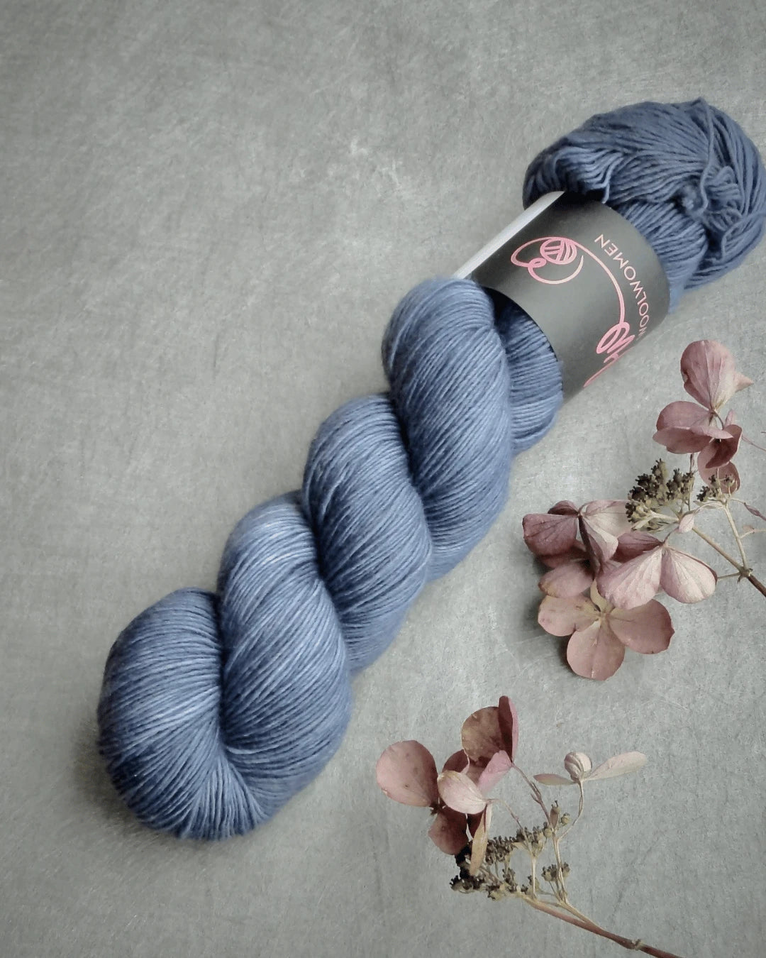 A skein of WoolWomen Merino Single yarn in soft blue rests on a gray surface next to dried pink and beige flowers. The partially visible label displays the product’s logo and text.
