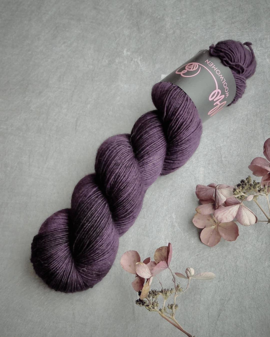 A skein of WoolWomen Merino Single in deep purple, with its label, rests on a gray surface beside dried pink and beige flowers.