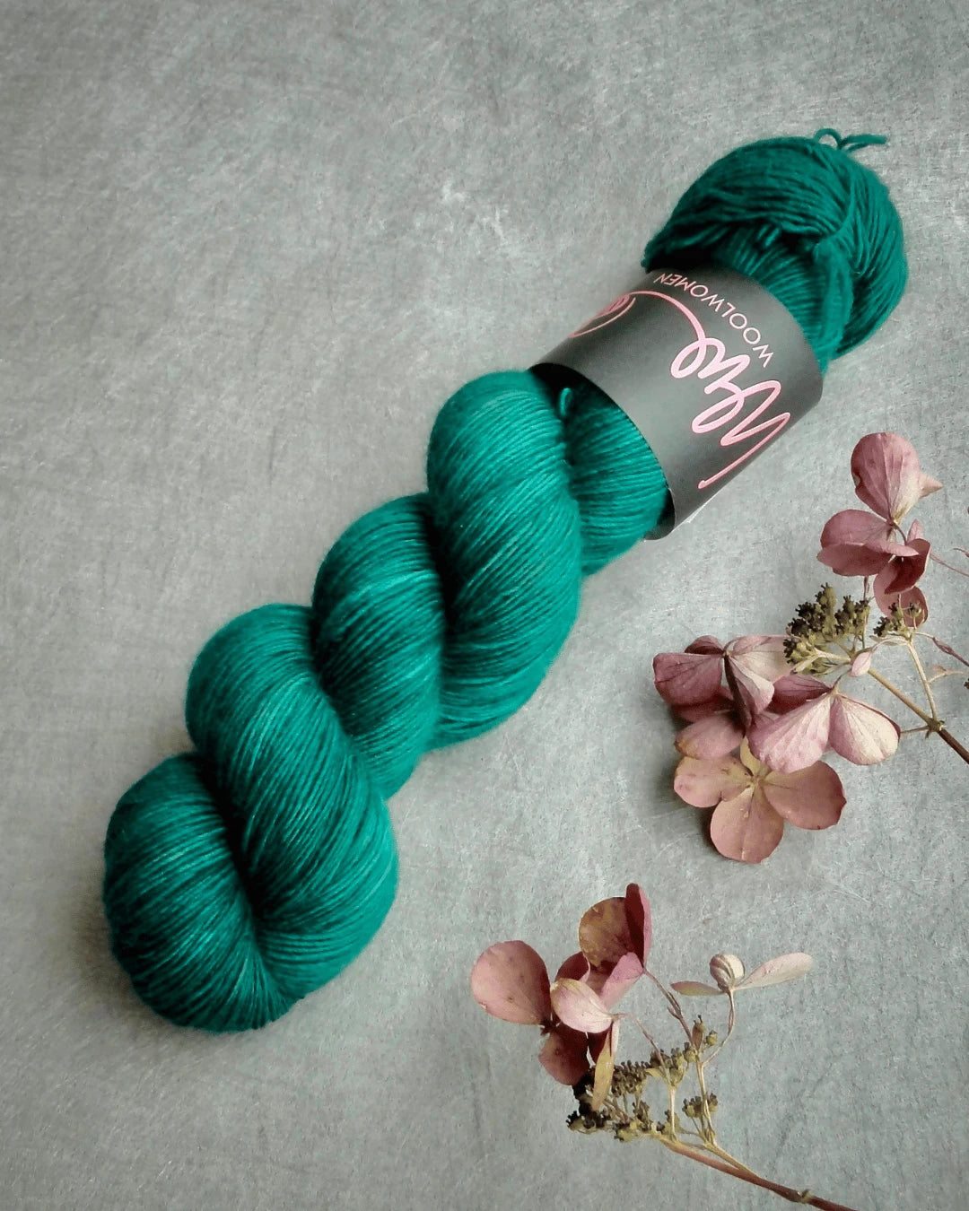 A skein of WoolWomen Merino Single, käsinvärjätty teal merinovilla yarn with a black label, rests on a gray surface beside dried pink and brown hydrangea flowers.