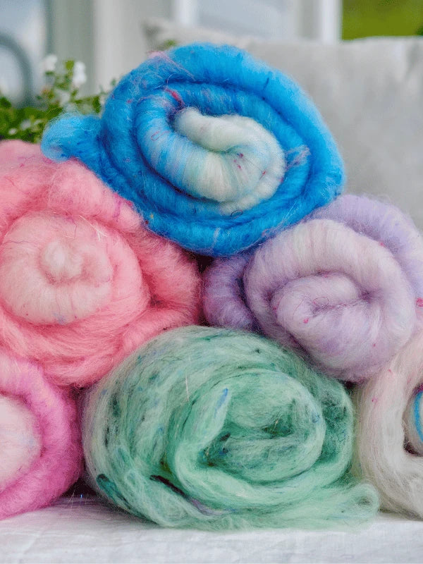 Six rolls of WoolWomen Art Batt - Corriedale, dyed in blue, pink, purple, and green, are stacked to showcase their soft texture. The blurred background features hints of greenery and a white cushion.