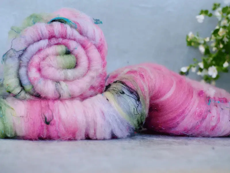 Two rolls of WoolWomen Art Batt -Cormo, featuring soft, colorful käsinvärjätty villa in shades of pink, green, and purple, rest on a light surface as a small blurred white flowering plant decorates the background.