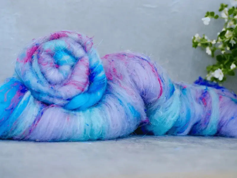 A bundle of WoolWomen Art Batt - Cormo kehräyskuitu in soft blue, purple, and pink hues is coiled on a light surface, accented by delicate white flowers in the background.