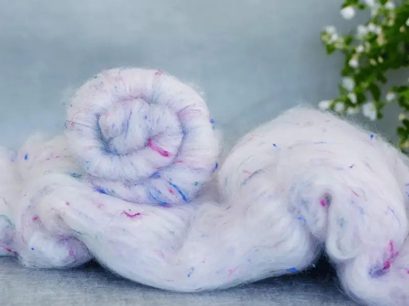 A close-up of the WoolWomen Art Batt -Polwarth, a soft, fluffy, käsinvärjätty villakuitu for spinning, with colorful blue, pink, and purple specks, partially rolled up against blurred green foliage and white flowers.