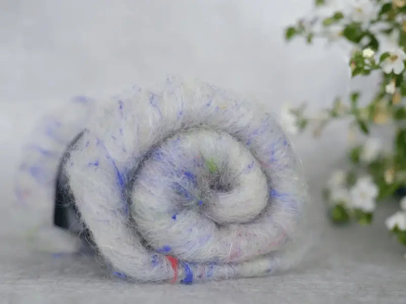 A close-up of WoolWomen Art Batt - Suomalainen villa, a soft, white wool with subtle blue and purple specks, rolled up on a light surface. Delicate white flowers with green leaves appear in the background on the right.
