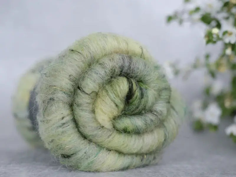 A close-up of WoolWomen Art Batt - Suomalainen villa shows a rolled bundle of soft, light green Finnish wool with subtle yellow and grey hues on a pale surface. Blurred white flowers and green leaves appear in the background.