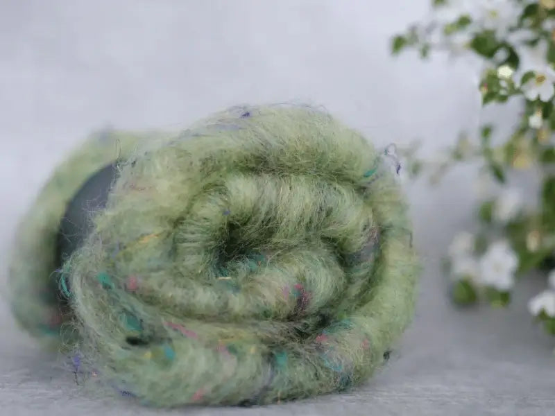 A close-up of a rolled WoolWomen Art Batt -Suomalainen villa in soft green with colorful flecks rests on a gray surface. Slightly blurred white flowers appear in the background, emphasizing its charm for kehräys projects.