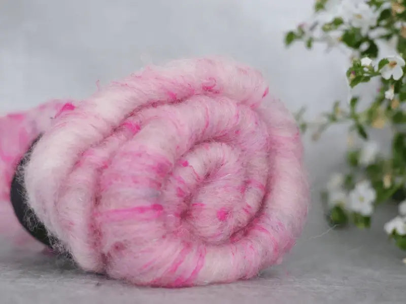 A close-up of a rolled bundle of WoolWomen Art Batt - Suomalainen villa, pink and white käsinvärjätty wool fiber, on a gray surface with blurred white flowers and green leaves in the background.