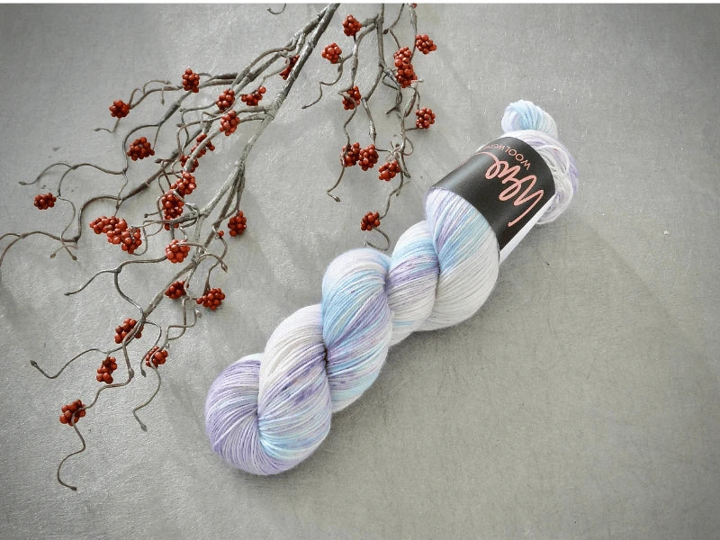 A skein of WoolWomen Merino Sock – Yhden Padan Ihme in pastel blue, purple, and white rests on a gray surface beside a branch with small red berries.