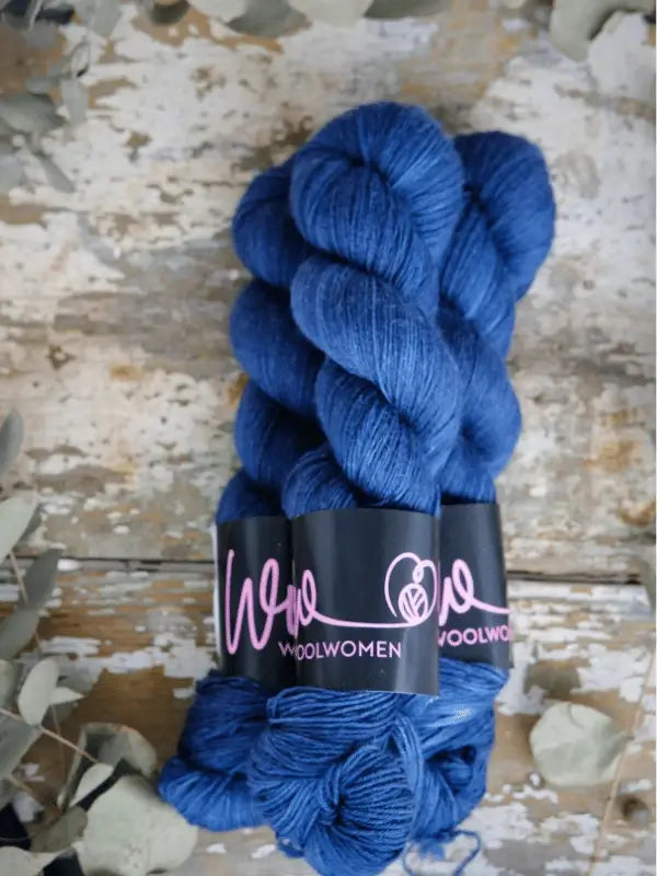 Two skeins of WoolWomen Merino Sock yarn in royal blue rest on a rustic wooden surface, surrounded by dried leaves and foliage.
