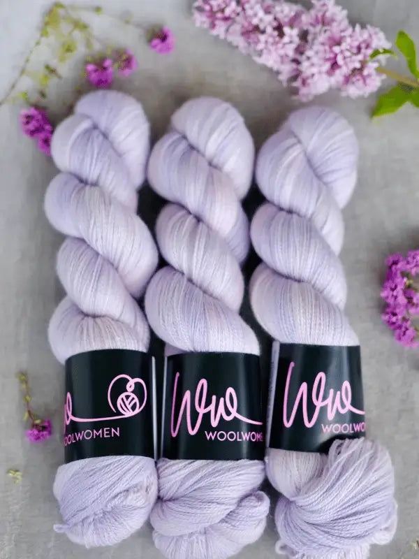 Three skeins of soft, pale lavender WoolWomen Merino Silk Lace yarn with black labels are arranged on a neutral surface, surrounded by clusters of purple and lilac flowers.
